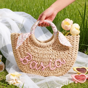May include: A woven straw handbag featuring a semi-circular shape and two round handles wrapped in a floral fabric. The front of the bag displays the name Basia in pink cursive yarn lettering. The bag is accompanied by a perfume bottle and sunglasses.