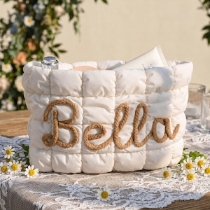 Könnte beinhalten: Ein weißer, gesteppter Stoffkorb mit dem Namen "Bella" in Seilbuchstaben. Der Korb enthält Schönheitsprodukte und steht auf einem mit Spitze bedeckten Tisch mit Gänseblümchen. Der Korb hat ein weiches, gepufftes Design.