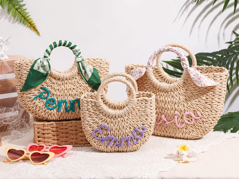 May include: Three woven straw handbags with rounded handles. The left bag features a green scarf and the name Penny in teal. The center bag displays the name Emma in purple. The right bag has a floral scarf and the name Asia in pink.