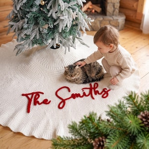 May include: A circular, cream-colored knitted Christmas tree skirt featuring the text "The Smiths" in bold, red, cursive lettering. The skirt has a ribbed texture and is positioned around the base of a flocked artificial Christmas tree.