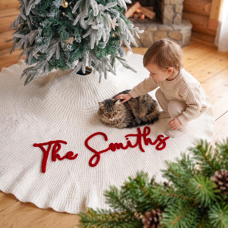 May include: A circular, cream-colored knitted Christmas tree skirt featuring the text "The Smiths" in bold, red, cursive lettering. The skirt has a ribbed texture and is positioned around the base of a flocked artificial Christmas tree.