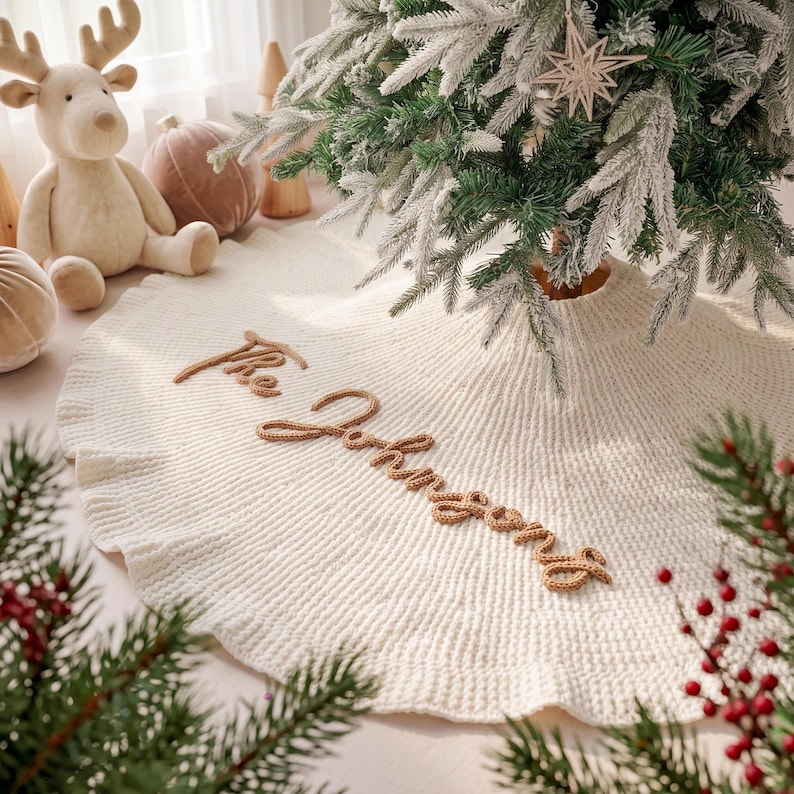 May include: A circular, cream-colored knitted Christmas tree skirt featuring the text "The Johnsons" embroidered in brown yarn. The skirt has a ribbed texture and is positioned beneath a flocked green Christmas tree with a star ornament.