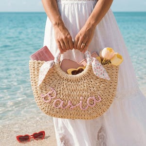 Può includere: Una borsa tote in paglia intrecciata con il nome Basia in corsivo rosa sul davanti. La borsa presenta due manici rotondi avvolti in un tessuto floreale con nastri abbinati. All'interno si trovano un portafoglio rosa, occhiali e tulipani gialli.