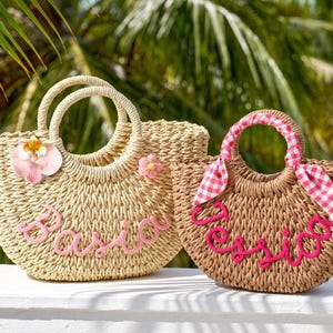 May include: Two woven straw tote bags with round handles. The larger bag is light beige with a pink flower, a pink orchid, and the name Basia in pink script. The smaller bag is tan with a pink and white gingham ribbon and the name Jessica in pink.