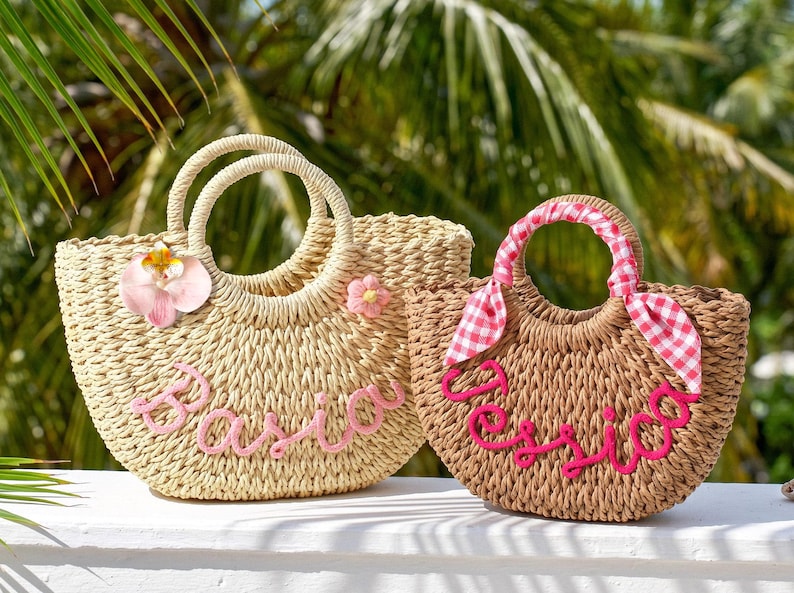 May include: Two woven straw tote bags with round handles. The larger bag is light beige with a pink flower, a pink orchid, and the name Basia in pink script. The smaller bag is tan with a pink and white gingham ribbon and the name Jessica in pink.
