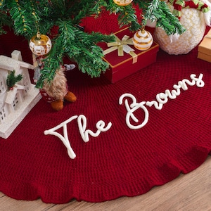 May include: A circular, deep red knitted Christmas tree skirt featuring the white embroidered text The Browns in a cursive script. The fabric has a ribbed texture and a ruffled edge, positioned beneath a green pine tree with ornaments.