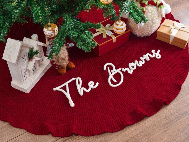 May include: A circular, deep red knitted Christmas tree skirt featuring the white embroidered text The Browns in a cursive script. The fabric has a ribbed texture and a ruffled edge, positioned beneath a green pine tree with ornaments.