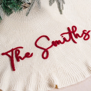 May include: A circular cream-colored knitted Christmas tree skirt featuring a textured fabric surface. The text "The Smiths" is embroidered in a bold, raised red script across the center of the fabric, creating a distinct contrast.