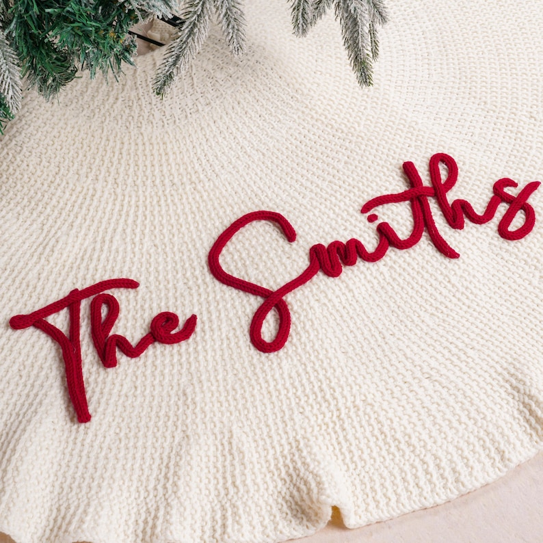 May include: A circular cream-colored knitted Christmas tree skirt featuring a textured fabric surface. The text "The Smiths" is embroidered in a bold, raised red script across the center of the fabric, creating a distinct contrast.