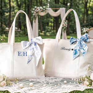 Pode incluir: Duas sacolas de lona creme com alças longas, decoradas com laços estampados em azul e branco. Uma sacola tem as iniciais "EH" bordadas em azul, a outra tem o nome "Beverly" com um símbolo de coração.