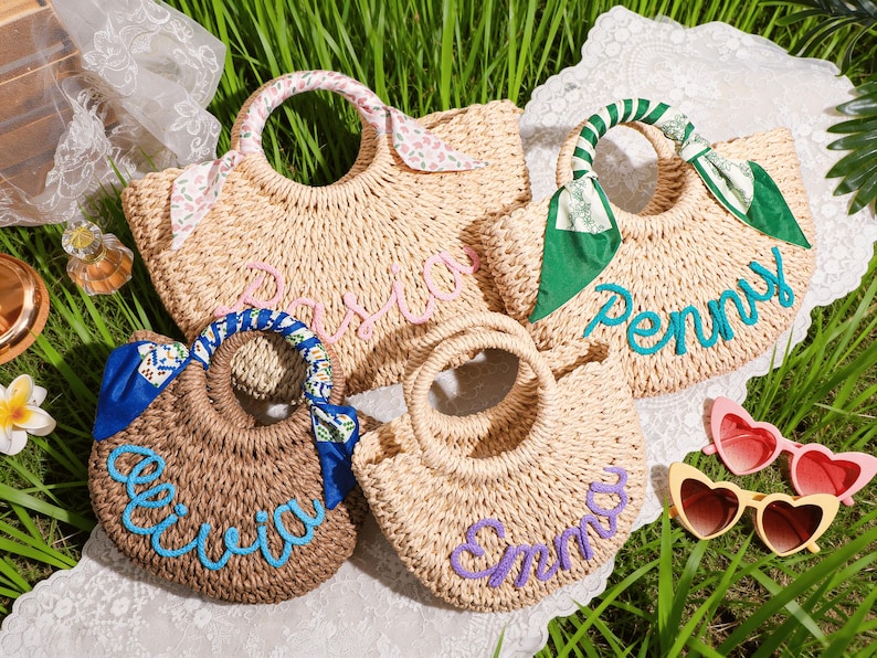 May include: Four woven straw handbags with round handles, each featuring a personalized name in cursive script. The bags are decorated with colorful fabric ribbons tied to the handles, displaying the names Pasia, Penny, Olivia, and Emma.