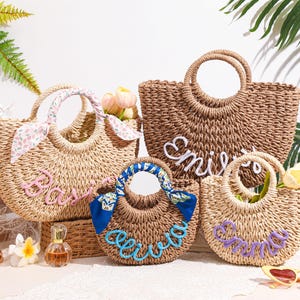 May include: Four woven straw tote bags with round handles, each featuring a personalized name in raised script lettering. One bag has a pink floral scarf on the handle, another has a blue patterned scarf, and the names are in pink, white, blue, and purple.