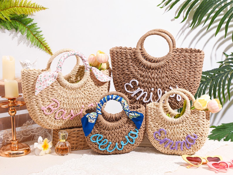 May include: Four woven straw tote bags with round handles, each featuring a personalized name in raised script lettering. One bag has a pink floral scarf on the handle, another has a blue patterned scarf, and the names are in pink, white, blue, and purple.