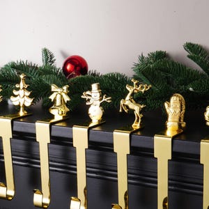 May include: Gold-coloured Christmas stocking holders featuring festive designs like a snowflake, Christmas tree, bell, snowman, reindeer, mitten, and wreath. They are arranged on a black surface with green garland and red baubles.