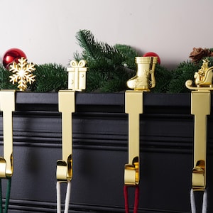 Può includere: Quattro porta calze natalizie color oro con disegni festivi: un fiocco di neve, un regalo, uno stivale e una slitta. Ogni supporto ha un anello per appendere ed è esposto su una superficie nera con verde.