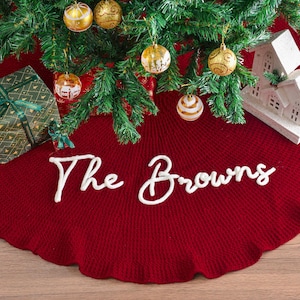 May include: A deep red circular knit Christmas tree skirt featuring a ruffled edge and the white cursive text The Browns centered on the fabric. The skirt is positioned beneath a green pine tree decorated with gold ornaments and a white house.