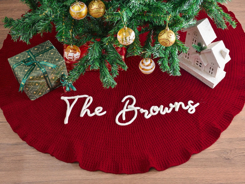 May include: A deep red circular knit Christmas tree skirt featuring a ruffled edge and the white cursive text The Browns centered on the fabric. The skirt is positioned beneath a green pine tree decorated with gold ornaments and a white house.