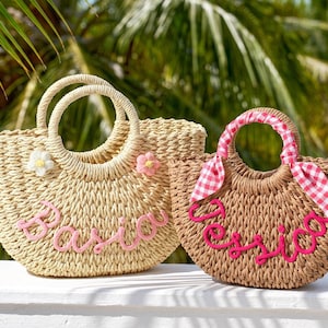 May include: Two woven straw handbags with round handles. The left bag is light beige with a white and pink flower, featuring the name Basia in pink script. The right bag is tan with a pink and white gingham ribbon and the name Jessica in pink.