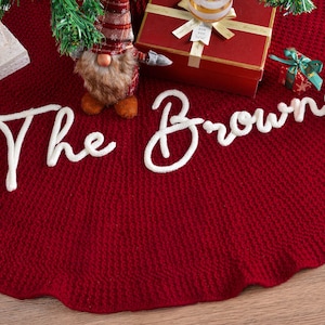 May include: A deep red circular Christmas tree skirt featuring a textured knit pattern and the white script text The Browns appliqued across the center. The skirt is displayed with a gnome figurine and gift boxes placed on its surface.