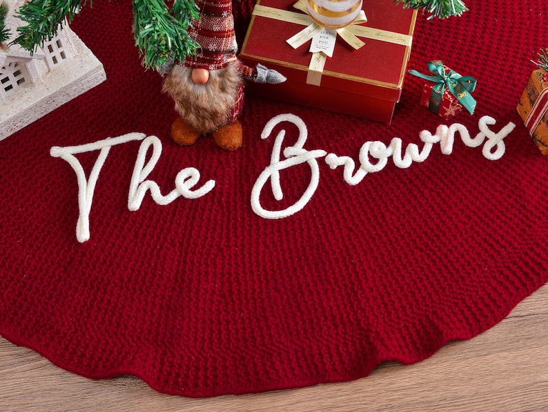 May include: A deep red circular Christmas tree skirt featuring a textured knit pattern and the white script text The Browns appliqued across the center. The skirt is displayed with a gnome figurine and gift boxes placed on its surface.