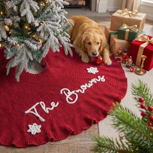 May include: A circular, deep red knitted Christmas tree skirt featuring white, cursive embroidered text that reads The Browns, accented by two white crocheted snowflake appliqu&eacute;s. The skirt is positioned under a flocked Christmas tree.