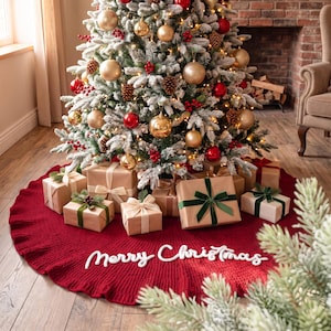May include: A circular, deep red knitted Christmas tree skirt with a ruffled edge and the white cursive text Merry Christmas embroidered on the front. The skirt is placed under a flocked Christmas tree decorated with gold and red ornaments.