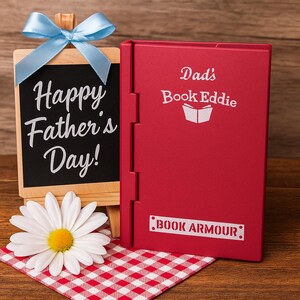 May include: A red book-shaped case with the text "Dad's Book Eddie" and "Book Armour". A small chalkboard with "Happy Father's Day!" is tied with a blue ribbon. A white daisy rests on a red and white checkered cloth.