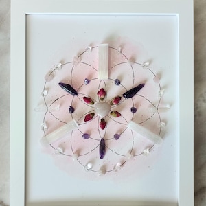 Puede incluir: Obra de arte enmarcada con un diseño geométrico que incorpora cristales, capullos de rosa secos y piedras de amatista. El diseño está sobre un fondo rosa de acuarela y enmarcado en blanco. Capullos de rosa y un cristal de selenita están dispersos.