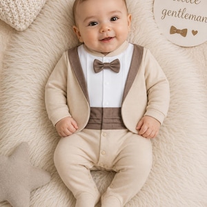 May include: A beige and white long-sleeved footed romper featuring a tuxedo-style design with a white front panel, a brown bowtie, a brown cummerbund-style waist detail, and a beige jacket-style overlay with brown lapels and snap closures.