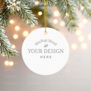 May include: A white, round Christmas ornament with the text "Mockup Mood YOUR DESIGN HERE" in gray. The ornament hangs from a gold string against a blurred background of lights and evergreen branches.