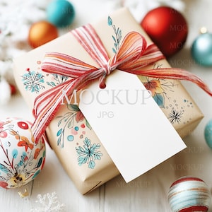 Puede incluir: Una caja de regalo envuelta con una cinta a rayas rojas y blancas y una etiqueta blanca en blanco. La caja está decorada con diseños florales y de copos de nieve. Adornos navideños rojos, azules y dorados rodean el regalo.