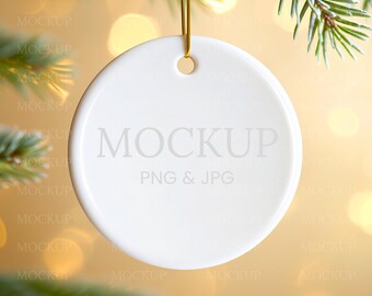 Könnte beinhalten: Ein weißer, runder Keramik-Weihnachtsbaumschmuck mit dem Aufdruck "MOCKUP" und "PNG & JPG". Er hängt an einem goldenen Faden, mit einem verschwommenen Hintergrund aus Tannenzweigen und Bokeh-Lichtern.