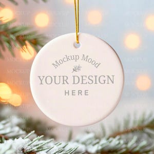 May include: A round, off-white Christmas ornament with the text "Mockup Mood YOUR DESIGN HERE" in gray. The ornament hangs from a gold string, with a blurred background of lights and evergreen branches.