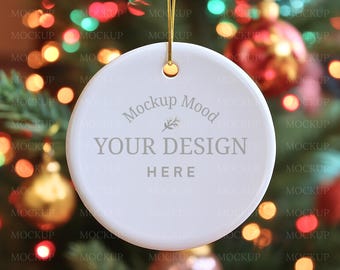 Könnte beinhalten: Ein weißer, runder Weihnachtsschmuck mit einem goldenen Faden zum Aufhängen. Der Schmuck trägt den Text "Mockup Mood" und "YOUR DESIGN HERE". Der Hintergrund ist ein verschwommenes Bild eines Weihnachtsbaums mit bunten Lichtern und Dekorationen.