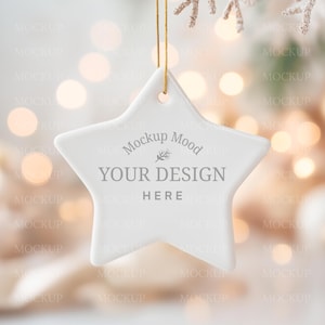 Könnte beinhalten: Ein weißer, sternförmiger Christbaumschmuck, an einem goldenen Faden aufgehängt. Auf dem Schmuck steht der Text "Mockup Mood YOUR DESIGN HERE". Der Hintergrund ist verschwommen mit warmen Bokeh-Lichtern, was eine festliche Stimmung erzeugt.