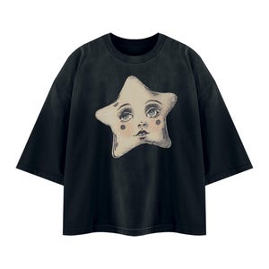 May include: A black short-sleeved t-shirt featuring a large, centered graphic of a cream-colored star with a detailed human face. The star illustration includes expressive eyes with long lashes, rosy cheeks, and a small mouth, rendered in a vintage style.