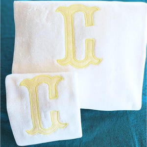Può includere: Un set di due asciugamani in spugna bianca, uno grande e uno piccolo, ciascuno caratterizzato da una grande lettera C ricamata al centro con filo giallo tenue, in un carattere decorativo con grazie e un bordo cucito in giallo.