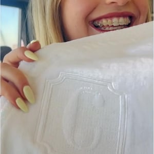 May include: White towel with an embroidered decorative border and a large letter "G" in the center. The towel is held up by a person with blonde hair and braces.