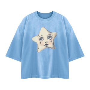 May include: A light blue short-sleeve t-shirt featuring a central graphic of a cream-colored star with a detailed human face. The star has large eyes with long eyelashes, defined eyebrows, a small nose, and pink-tinted cheeks on a solid blue background.