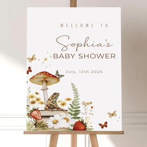 May include: A white sign on a wooden easel with the text "Welcome to Sophia's Baby Shower, July, 12th 2026." The sign features watercolor illustrations of a mushroom, frog, strawberries, daisies, and butterflies.