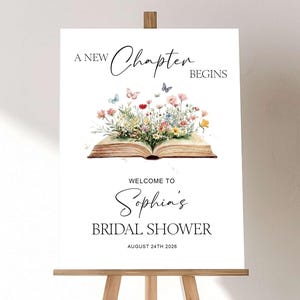 May include: A white rectangular sign on a wooden easel featuring the text A NEW CHAPTER BEGINS, WELCOME TO Sophia's BRIDAL SHOWER, and AUGUST 24TH 2026. An illustration of an open book with colorful wildflowers and small butterflies emerging from the pages.