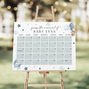 May include: A baby shower calendar sign on an easel. The sign is white with a blue and white design, including clouds, stars, and a hot air balloon with a teddy bear. The text reads "guess the arrival of BABY EVAN" and includes a calendar.