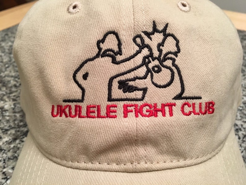 May include: A beige baseball cap with a black embroidered illustration of two figures fighting with ukuleles. The text "Ukulele Fight Club" is embroidered in red below the illustration.