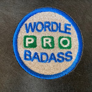 Puede incluir: Un parche bordado blanco y azul con el texto "WORDLE PRO BADASS" en negro. Las letras "PRO" están en cuadrados verdes.