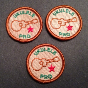 Puede incluir: Tres parches bordados de color marrón y beige con el texto "Ukulele Pro" y una estrella roja. Cada parche presenta un ukelele.