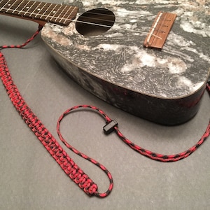May include: A red and black braided ukulele strap with black plastic clips. The strap is made of paracord and is designed to be durable and comfortable.
