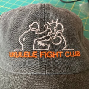 May include: A black baseball cap with an embroidered design of two figures playing ukuleles. The text "Ukulele Fight Club" is embroidered in orange below the design.
