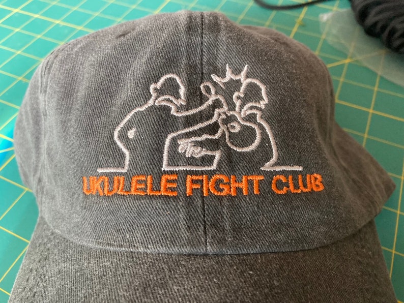 May include: A black baseball cap with an embroidered design of two figures playing ukuleles. The text "Ukulele Fight Club" is embroidered in orange below the design.