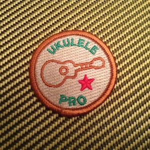 May include: A brown and orange embroidered patch with the text "Ukulele Pro" in green lettering. A white ukulele with a red star is in the center of the patch.