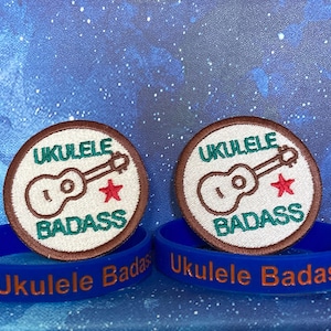 Op de afbeelding: Twee bruine en witte geborduurde patches met een ukelele en de tekst "Ukulele Badass". Twee blauwe siliconen armbanden met de tekst "Ukulele Badass" in oranje.